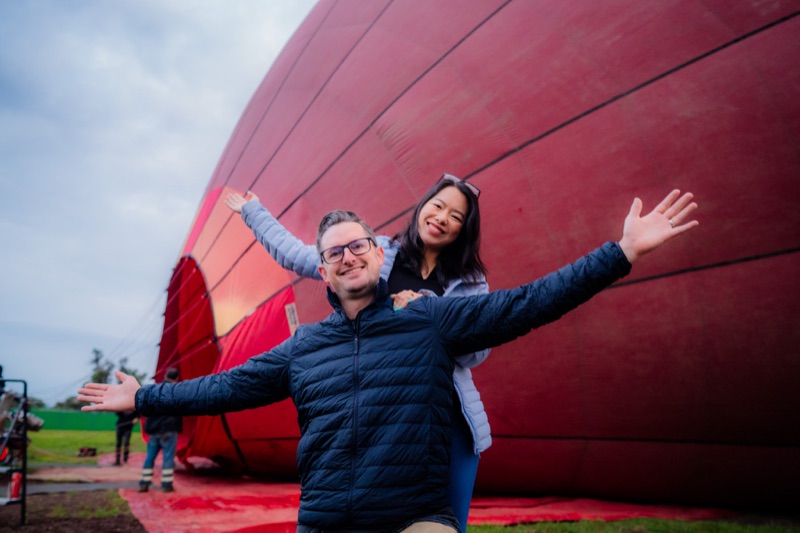 Pareja con los brazos abiertos frente a un globo aerostático rojo mientras se infla en el campo