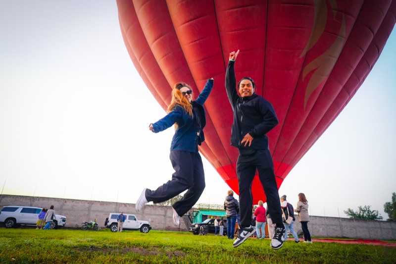 Pareja saltando de emoción frente a un globo aerostático rojo en el campo de despegue de Teotihuacán