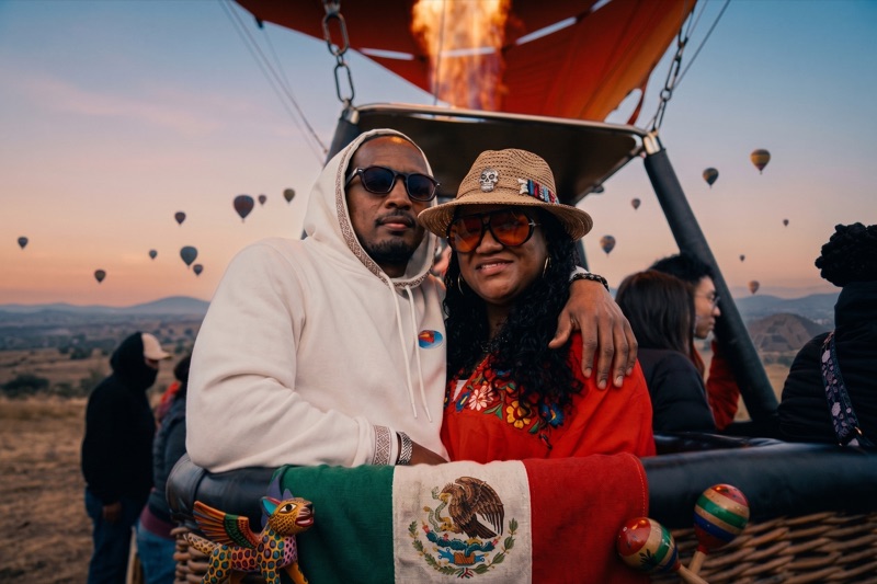 Pareja en la canasta de un globo aerostático al amanecer sobre Teotihuacán, con decenas de globos en el cielo y decoración mexicana